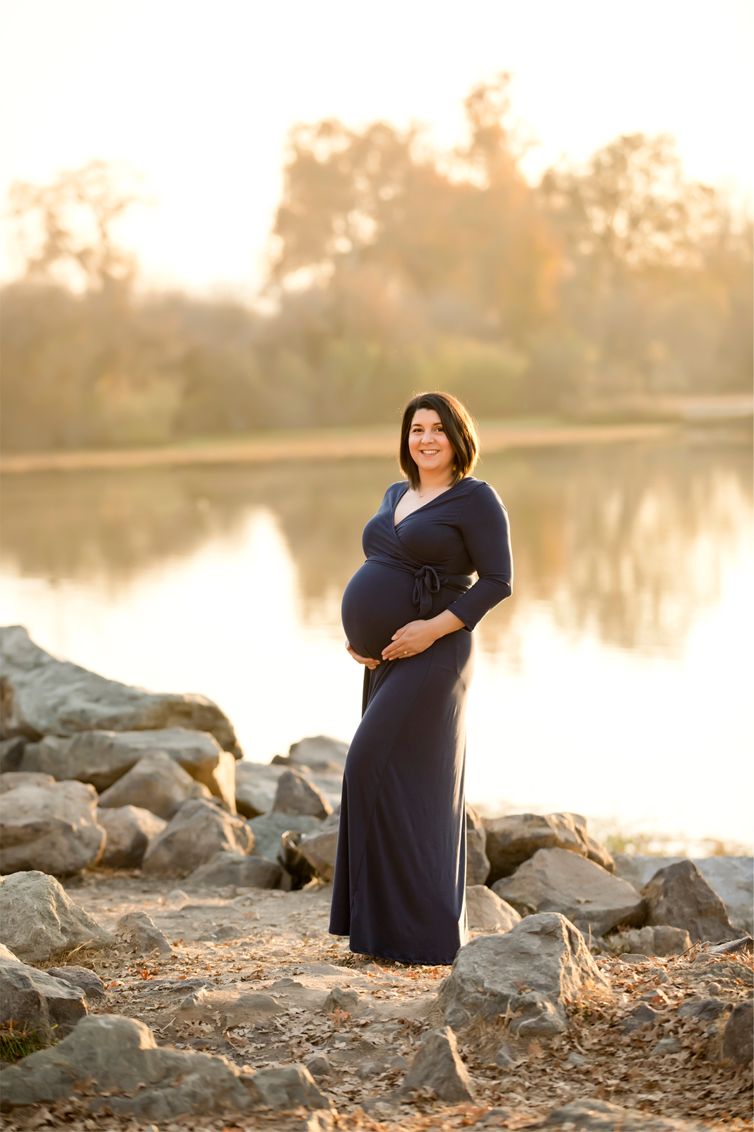 Pregnant woman in navy long sleeve dress standing in front of a lake at sunset