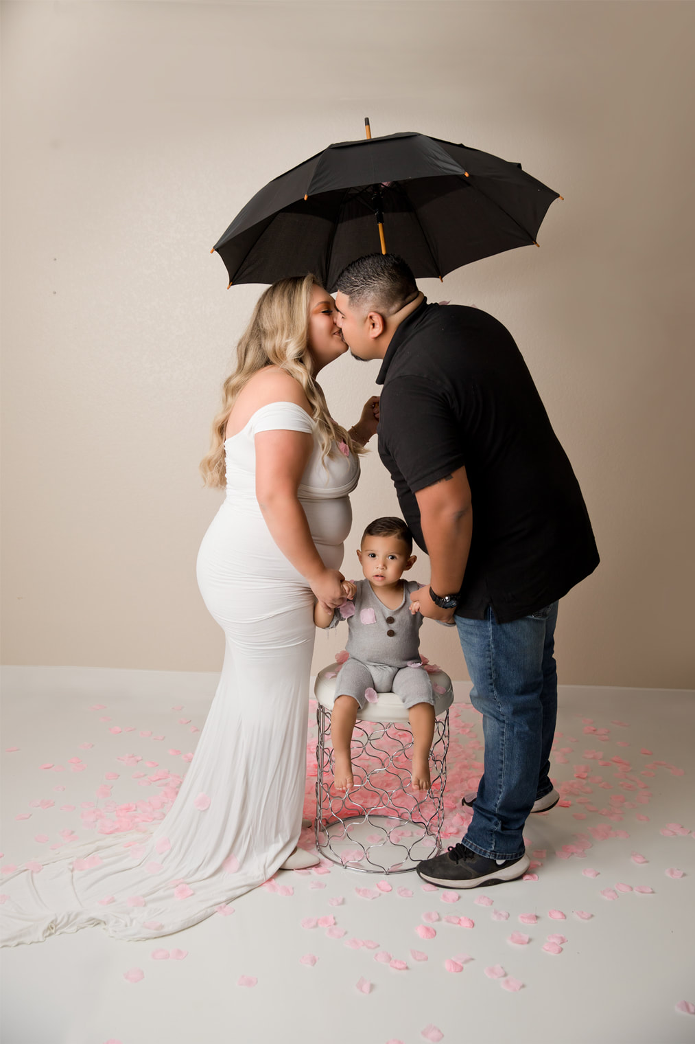 Couple kissing with umbrella overhead, pink confetti on ground, little boy on stool between them