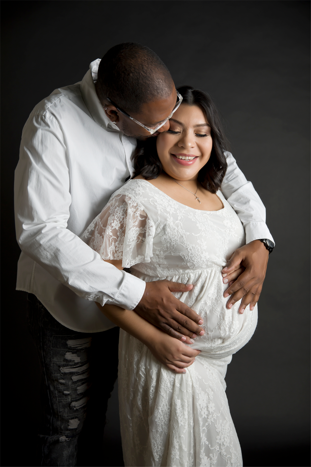 Couple, man kissing woman's head, both holding her pregnant belly, dark gray backgrounde
