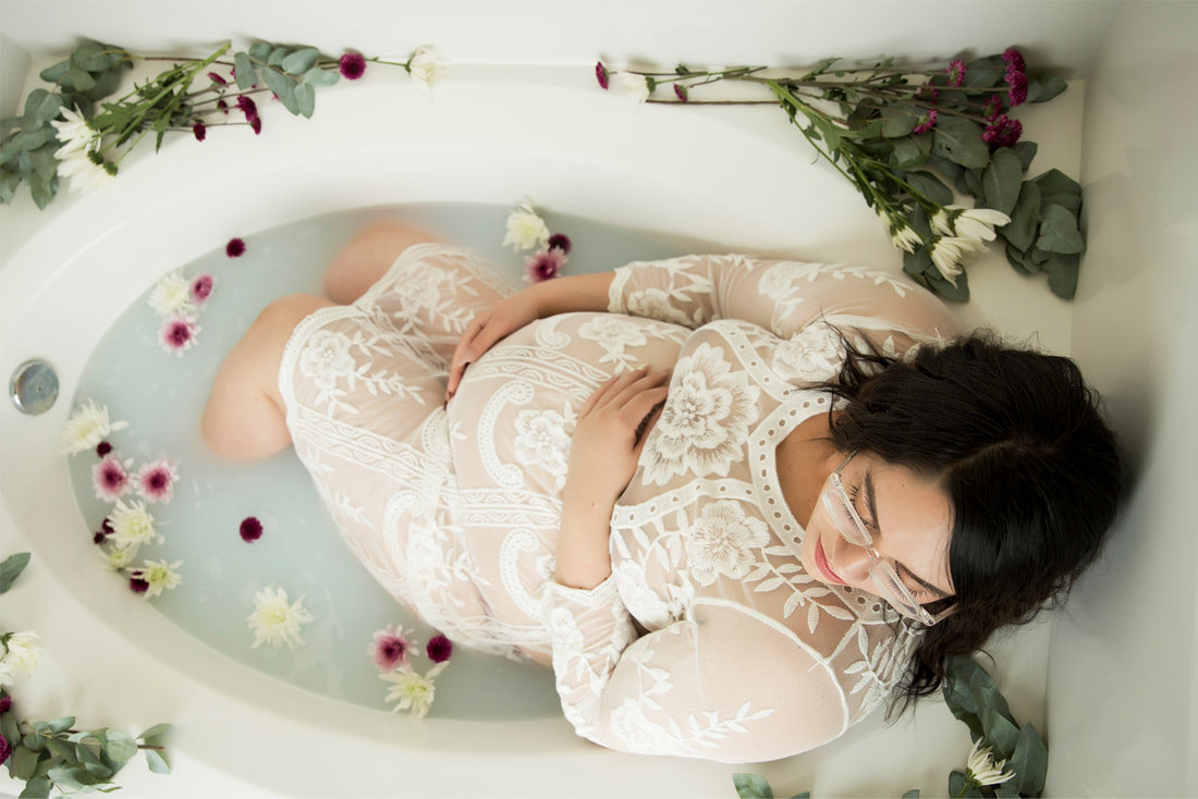 Woman in white lace dress laying in white tub with milk and pink and white flowers, holding pregnant belly.