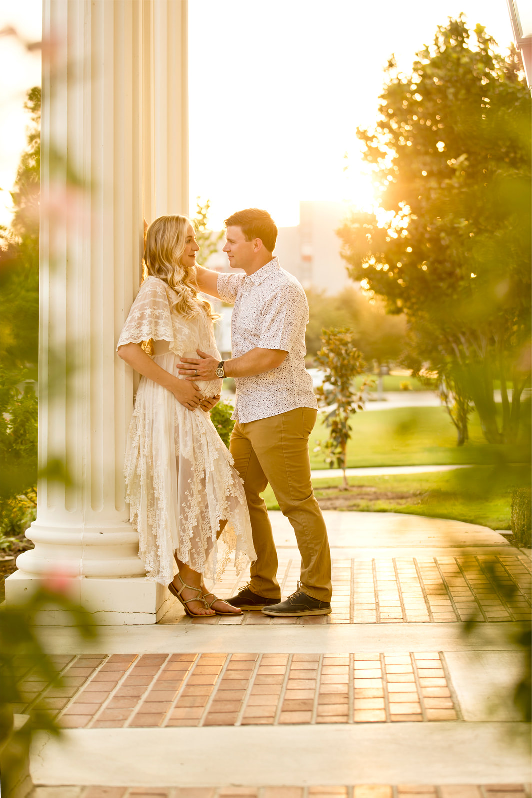 Man and pregnant woman leaning againt a pillar, sunset and trees behind them