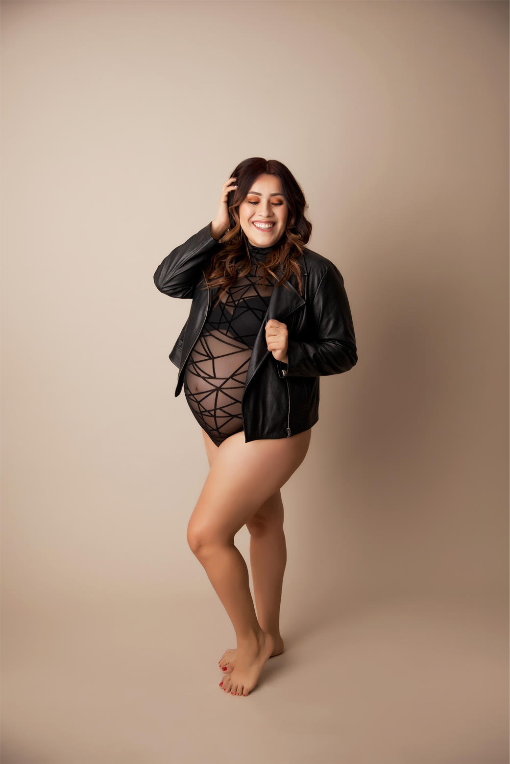 Pregnant woman in geometric mesh black onesie, black leather jacket, neutral background