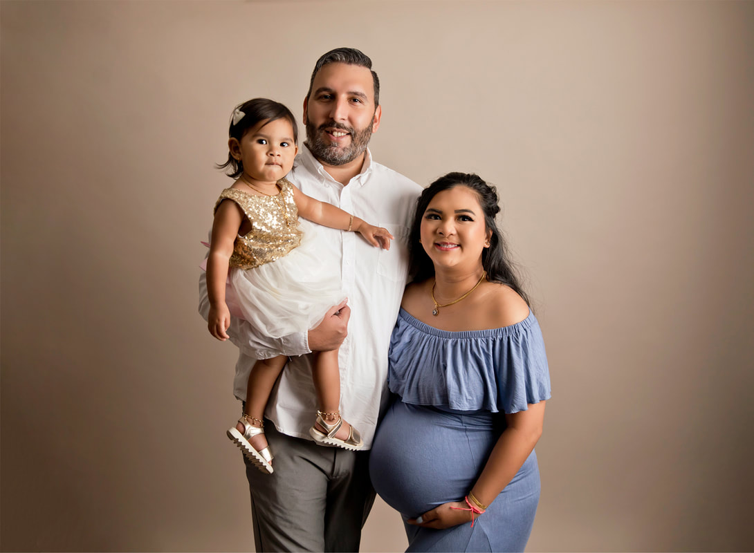  Woman holding pregnant belly in blue dress, man holding little girl.Tan background