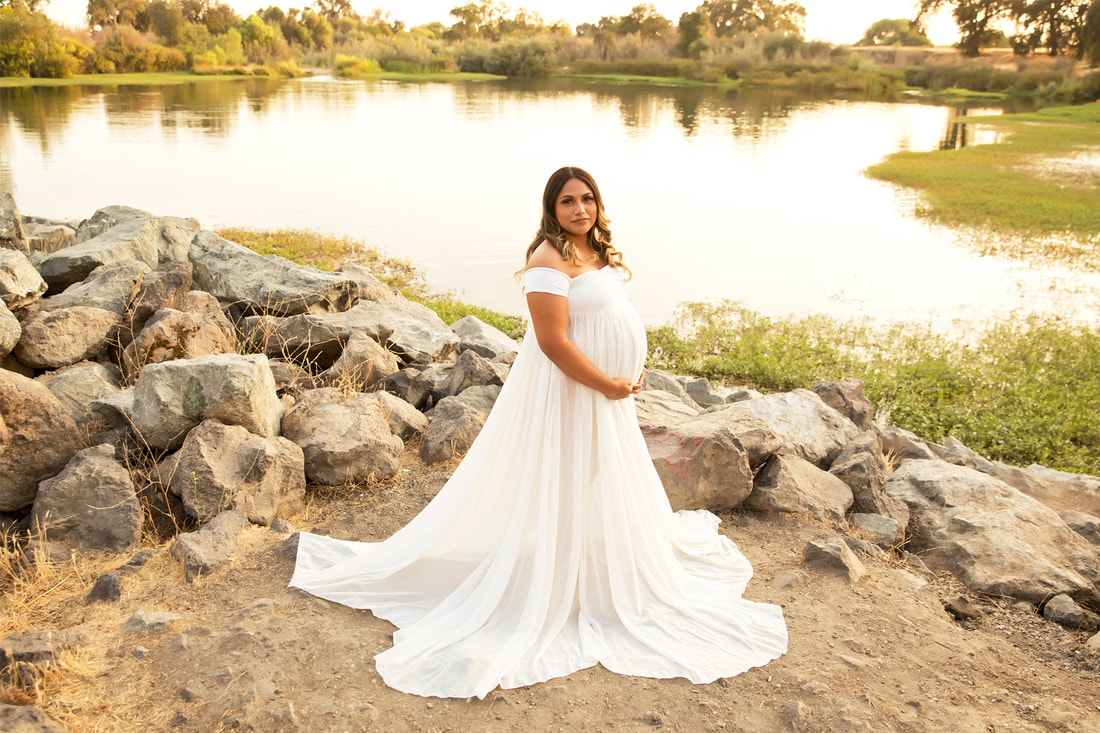 Pregnant woman in flowy white dress standing in front of a lake