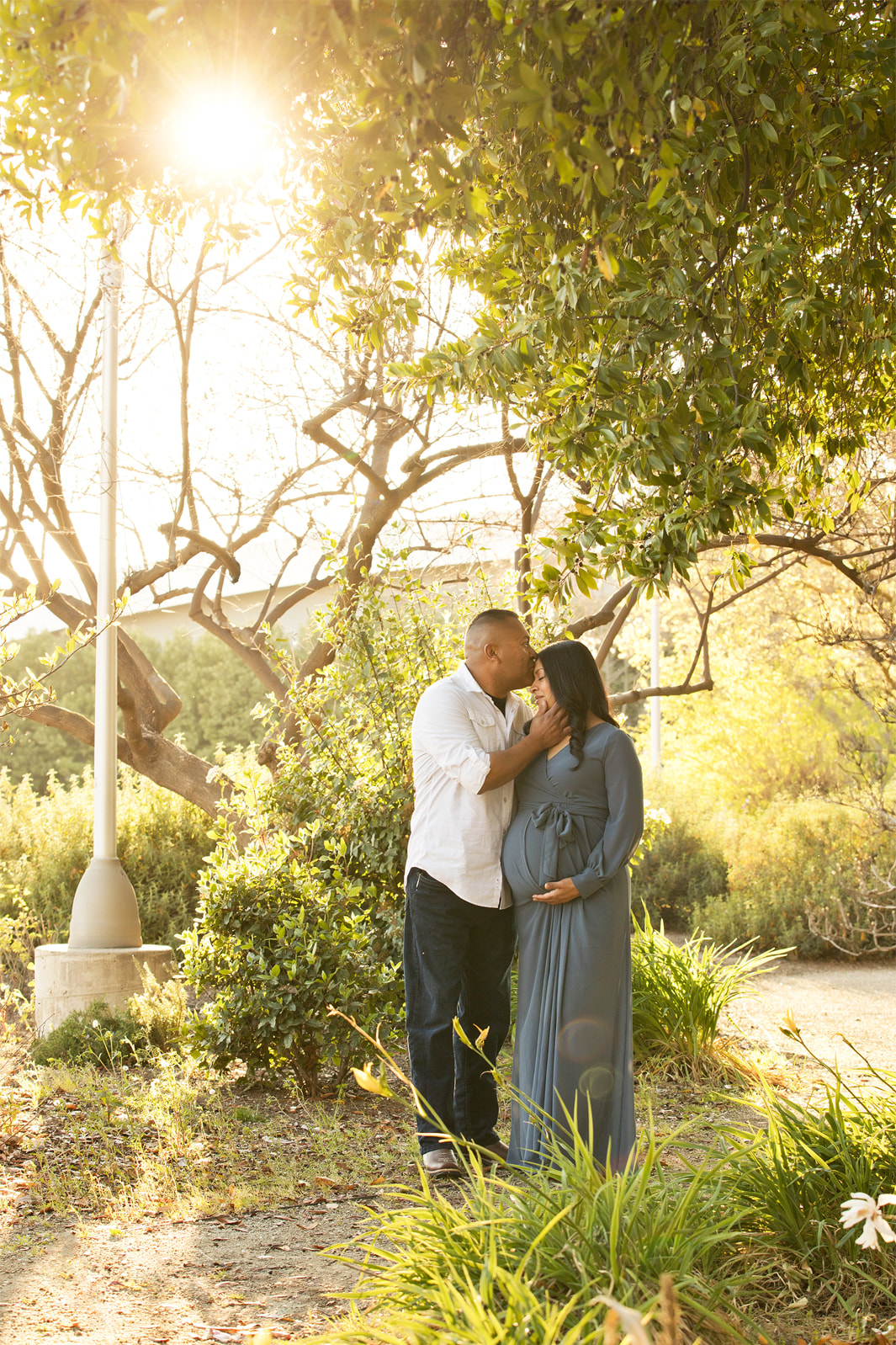 Man kissing pregnant woman's forehead, standing in greenery
