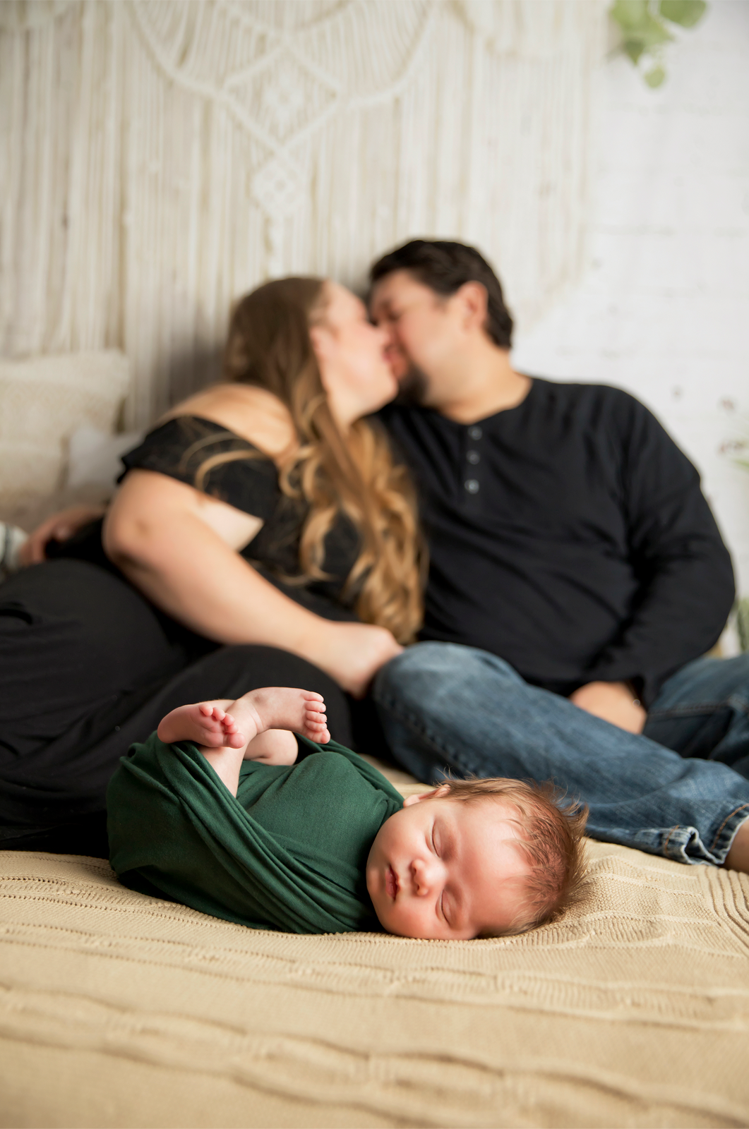 Sleeping baby boy in green on bed in front of woman and man kissing in front of boho background. 