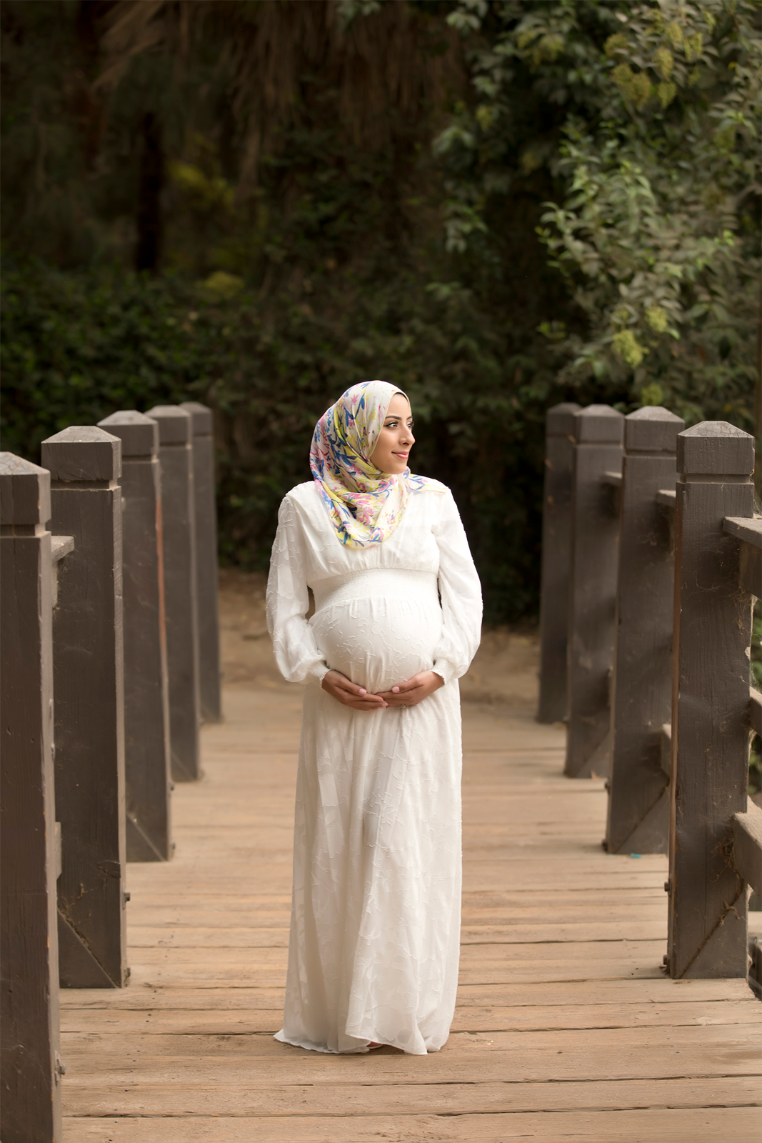 Woman in white dress and hijab holding her belly standing on a bridge