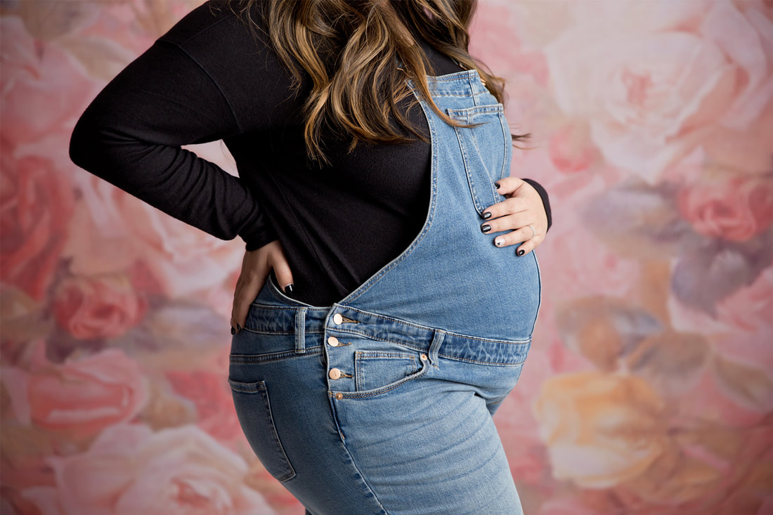 Pregnant woman in overalls and black long sleeve, her face isnt showing, holding belly, floral pink backdrop