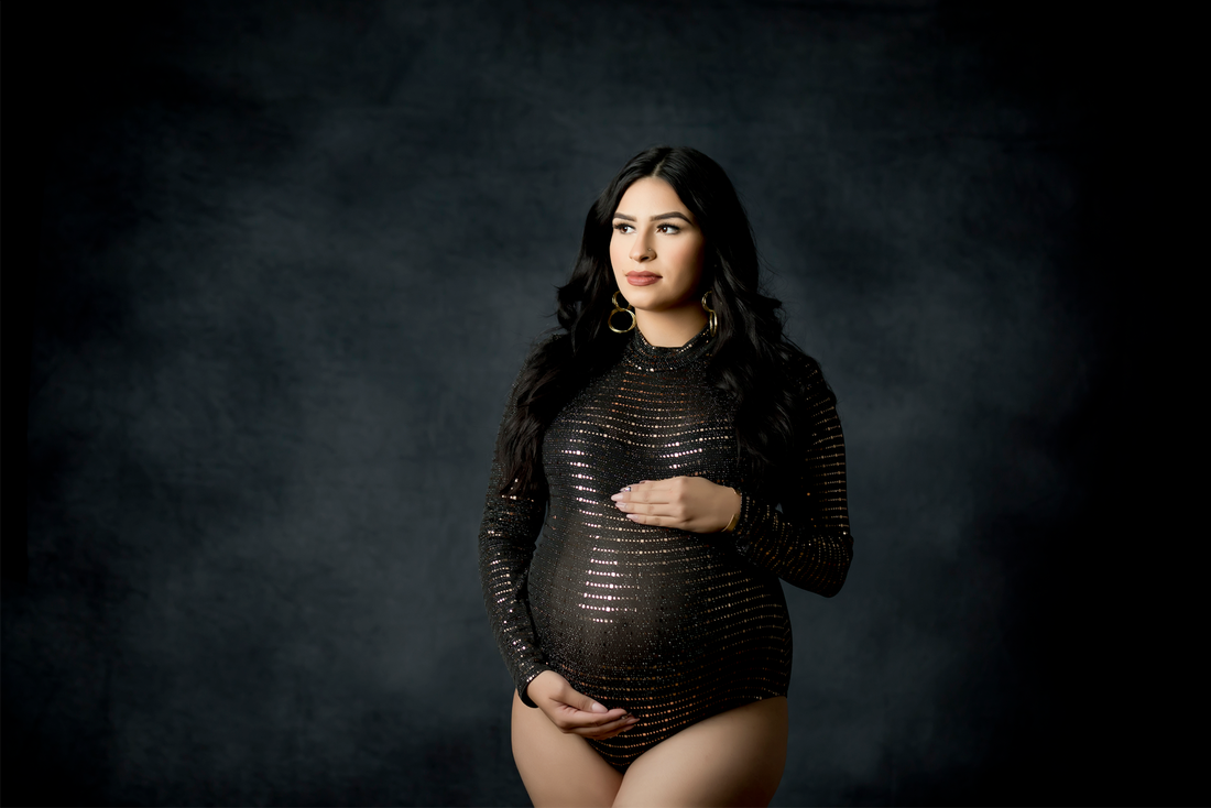 Woman in a black onesie holding pregnant belly, standing in front of dark blue backdrop