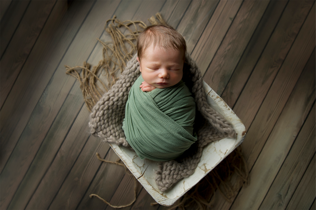 Baby wrapped in green swaddle laying in a white bowl on wood floor.
