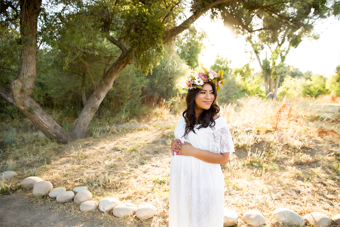 Woman in white dress and flower crown looking off into the distance holding her pregnant belly, field and trees behind her