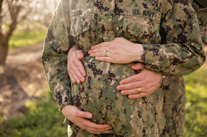 Two pairs of hands holding a pregnant belly, both wearing military uniform