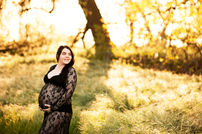 Pregnant woman looking into the distance, wearing a black lace dress and sun soaked greenery behind her