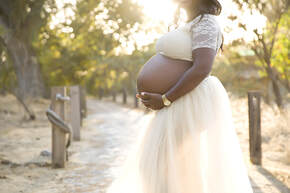 Standing woman holding her pregnant belly, wearing a two piece cream outfit in an orchard