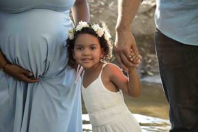 Man and woman holding hands with little girl, wearing a flower crown and white dress. The woman is holding her pregnant belly