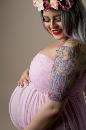 Woman in pink dress holding pregnant belly, wearing a flower crown, against tan backdrop