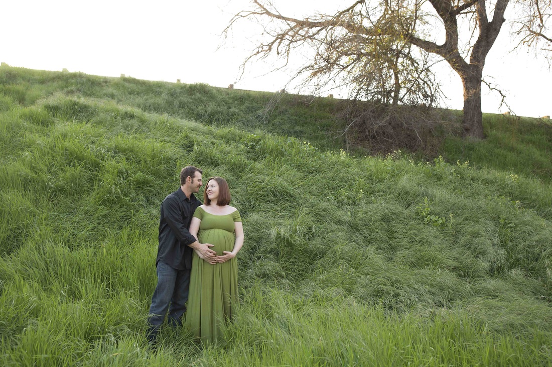 Man holding pregnant woman's belly, she is in a green dress, he is in a shirt and pants. Lush greenery behind them