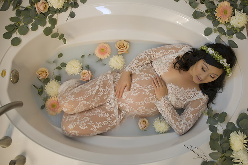 Woman in white lace dress laying in white tub with milk and yellow flowers, holding pregnant belly.