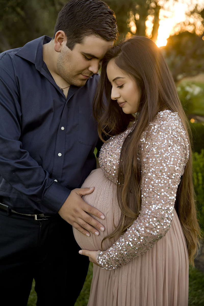 Man in navy shirt holding woman's pregnant belly, she is wearing a sparkly dress, sun shining behind them.